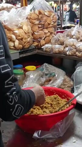 The video shows a street food vendor in a bustling market.  A person with a black long-sleeve shirt is using their hands to fill small, round puri shells with a yellow chickpea mixture from a large red bowl.  The filled puris are then placed into clear plastic bags alongside other pre-filled bags.  Behind the vendor, there are numerous bags filled with unfilled puris.  The market is crowded with people, and there are various stalls selling different items. The environment appears to be a lively and busy market in an outdoor or semi-outdoor setting.