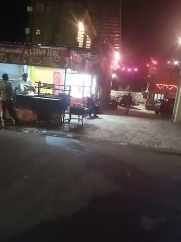 The video shows a nighttime street scene. There's a food stall labeled "CHOMA ZONE" to the left, with a man standing behind the counter, possibly cooking or serving. Several people are seated nearby. The street is paved with both dark asphalt and lighter-colored stones. Further down the street, several vehicles, including a blue-lit car, are visible. String lights decorate the area. The video appears to be taken from inside a moving car, with the windshield reflecting light.