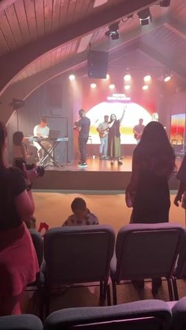 A worship band performs on a stage under a wooden A-frame ceiling. The stage is lit with warm, colorful lights, and a large circular graphic with a sunset design is projected behind the band. The band consists of a keyboard player on the left, a violinist and singer in the center, and two guitarists on the right. Several other band members are visible further back. In the foreground, there are rows of empty chairs. A child is sitting on the floor in front of the chairs, facing away from the stage. Several people are standing in the aisle between the chairs and the stage, facing the performers. One person in the foreground is holding a camera and appears to be filming the performance. The overall atmosphere appears to be a religious service or gathering.