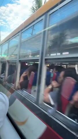 The video appears to be shot from inside a moving bus. The camera pans across the side of the bus, showing passengers through the windows. Outside the bus, a street scene unfolds with buildings, power lines, and parked motorcycles. The bus itself is visible, with its windows and part of its exterior. Some passengers are seen leaning out of the windows. The environment appears to be a tropical or sub-tropical urban setting, possibly in Southeast Asia, judging by the architecture and vegetation. The sky is visible and appears partly cloudy. The overall impression is a snapshot of daily life and travel within a bustling town or city.