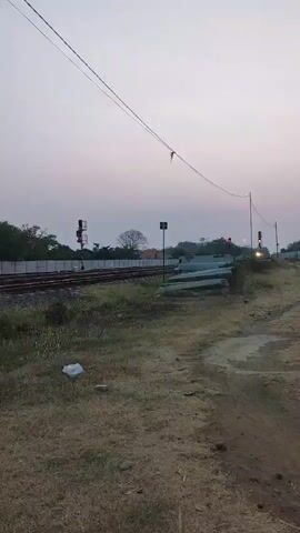 The video captures a railway line at dusk, with a signal light showing red, indicating a stop. The sky is a soft gradient of pink and pale blue. In the foreground, dry grass covers the ground, with a small white object, possibly litter, visible. A dirt path runs along the right side of the frame. Several concrete pipes are stacked near the railway tracks. Overhead, power lines stretch across the sky. The overall atmosphere is quiet and still.