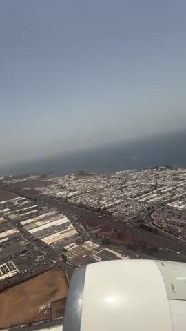 The video shows an aerial view from an airplane window during the daytime. The plane's white wing is visible in the foreground. Below, the landscape consists of a coastal city with mostly white buildings near the ocean, an industrial area featuring large rectangular buildings and parking lots, and stretches of brown, dry land. Several roads and roundabouts are seen connecting the different areas. The sea is calm and light blue, with a slightly hazy horizon. The sky above is clear and light blue. There are no people visible in the frame.