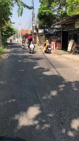 The video shows a street scene from the perspective of someone riding a motorcycle. In the foreground, the road surface is visible, with shadows cast by trees. Further down the road, three other motorcycles are traveling in the same direction. The first motorcycle, closest to the viewer, is white and ridden by two people. The second motorcycle is black and also has two riders. The third motorcycle is also black and appears to be carrying a single rider. The road is lined with buildings, some of which appear to be shops or homes. The weather is sunny.