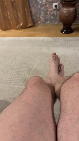 The video shows a first-person perspective of a person's legs and feet. The person appears to be sitting on a carpet with a geometric pattern. Their bare feet are visible, and their legs are crossed. The background is blurred but shows a wall with floral wallpaper, a curtain, and a vase. The lighting is dim, suggesting an indoor setting. The person moves their legs and feet slightly throughout the short clip.