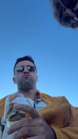 The video begins with a close-up shot of a person wearing sunglasses and an orange sweater, holding a glass of white wine. Their head is tilted back, looking up at the sky. As the video progresses, other people lean into the frame, also looking down at the camera. One person is wearing a red beret, and another is making a peace sign with their fingers. The camera angle is from below, looking up at the group. The background appears to be a clear blue sky. The individuals are interacting and seem to be in a jovial mood, possibly taking a group selfie or video.