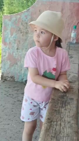 A young child wearing a pink t-shirt, white shorts with a teddy bear pattern, and a straw hat is standing next to a wooden plank. The child is looking down at their hands and holding a small rock. The background shows a textured wall with pink and blue paint, greenery, and a bottle of water on a shelf.