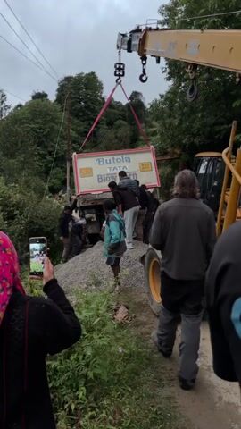 A crane is lifting a large white sign that says "Butola TATA Blowhorn" with a speed limit sign of 40 KM and "UK15CA 8555" on it. Below the sign is the back of a truck that appears to be stuck in gravel. Several men are gathered around the truck, some looking at the sign, others appearing to be helping or observing. One person in the foreground is holding up a phone to record the scene. The environment appears to be outdoors with trees and foliage in the background and a cloudy sky.