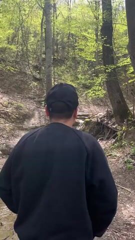 A young man in a black sweatshirt and black baseball cap stands by a shallow stream in a forest. He turns and looks at the water flowing over the small stone steps, then gestures towards it with his hand before walking away out of view. The stream flows through the forest, bordered by bare earth and fallen logs. Trees surround the scene, their leaves newly unfurled in the spring sunshine.