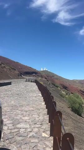 This media file captures a scenic view of a volcanic landscape under a clear blue sky. In the foreground, a cobblestone path lined with a rustic wooden fence leads towards a winding road. The path is empty, suggesting a quiet moment. Beyond the road, a steep, reddish-brown mountain rises, sparsely vegetated with patches of green shrubbery. Atop the mountain, several white observatory domes are visible, hinting at a scientific or astronomical facility.

The time of day appears to be midday, given the bright sunlight casting shadows and the intensity of the blue sky, which is dotted with a few wispy white clouds. The weather seems to be warm and clear, ideal for outdoor exploration. The landscape's reddish hue strongly suggests a volcanic origin, and the presence of observatories indicates a location with high altitude and clear atmospheric conditions, often sought for astronomical research. The scene evokes a sense of tranquility and awe, with the stark beauty of the natural landscape juxtaposed with the man-made structures of the observatory.

No people are visible in the frame, focusing the viewer's attention entirely on the natural and architectural elements of the scene. The location is identified as La Orotava, Spain, which is known for its volcanic terrain and high peaks like Mount Teide, a fitting setting for observatories. There is no visible text in the media file.