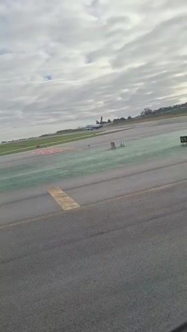 The video shows an airport runway on a cloudy day. In the distance, an airplane can be seen moving along the runway. The runway is marked with various lines and symbols. The foreground is out of focus, but the runway markings and the plane in the background are relatively clear. The environment is an airport with a runway and grassy areas surrounding it.