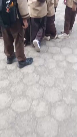 The video shows a group of children in uniform, likely scouts, standing in a line outdoors on a paved area. The children are wearing beige shirts with brown pants and red and white neckerchiefs. Some of the children are wearing head coverings. In the foreground, one child in a black cap and beige uniform is looking at a table. There is a woman in a yellow headscarf and patterned top standing behind the table, holding a microphone and a phone. She appears to be speaking to the children. On the table, there are bottles of water and what appears to be a map or game board. Other children and adults are visible in the background, with some forming a crowd. The environment appears to be an outdoor event or school activity.