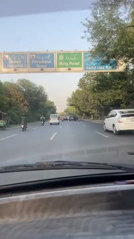 The video shows a first-person view from inside a car driving on a multi-lane road in Rasoolpur, Pakistan. The sky is bright and clear, suggesting daytime.

In the foreground, the car's dashboard and windshield wipers are visible. Ahead, the road stretches forward with two visible lanes in the direction of travel, marked by white dashed lines. Several vehicles are on the road, including a white Toyota Prius sedan on the right, a white van, and several darker colored cars further ahead. On the left lane, two motorcyclists are also seen. Lush green trees line both sides of the road, creating a somewhat enclosed and shaded environment.

Prominently displayed above the road are large overhead signs with destinations in both Urdu and English. From left to right, the signs indicate:
- Partially visible text, likely "Jinnah Hospital" (جناح ہسپتال)
- "Ferozepur Road" (فیروز پور روڈ)
- "Ring Road" (رنگ روڈ)
- "Johar Town" (جوہر ٹاؤن)
- "Abdul Haq Road" (عبدالحق روڈ)

The signs have green and blue backgrounds with white text and directional arrows. The overall scene depicts typical traffic flow on a well-maintained road in Pakistan.