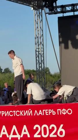 A scene unfolds on an outdoor stage featuring several people in white shirts and dark pants or skirts. Two individuals in purple shirts stand center stage, one speaking into a microphone. In front of them, several figures are kneeling or bent over, seemingly attending to someone lying on the floor. A red banner at the front of the stage reads "ТУРИСТСКИЙ ЛАГЕРЬ ПФО АДА 2026". Structural elements of the stage framework are visible overhead. A partial view of a flag is visible to the right. In the background, foliage and other people are faintly visible. The sky is a clear blue.