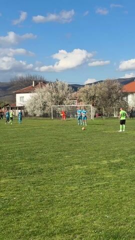 The video captures a soccer match in progress. Players in light blue and neon green jerseys are on the field, the blue team defending their goal and the green team attempting a free kick. The player in the green jersey takes the shot, the ball flies towards the goal, and the goalkeeper in an orange jersey attempts to block. The environment includes a grassy field, a white goal, houses with red tile roofs, trees with white blossoms, and distant mountains against a backdrop of blue sky dotted with white clouds.