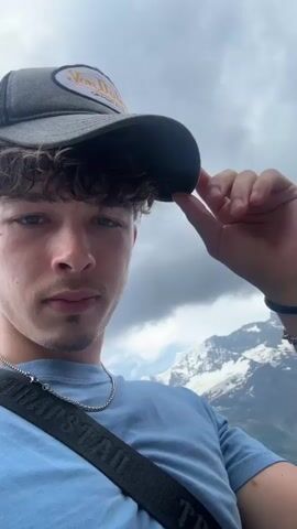 The video opens with a close-up selfie of a young man wearing a grey trucker hat and a light blue t-shirt. He is holding the brim of his hat with his right hand, and a silver chain is visible around his neck. A black strap with white lettering, possibly from a bag, crosses his chest. His expression is neutral, and he appears to be looking directly at the camera.

The camera then pans to the right, revealing a breathtaking mountain landscape. The background features towering, snow-capped mountains under a cloudy sky. Below the mountains, a charming village with traditional Swiss chalets is nestled on a hillside. The architecture of the houses, with their wooden facades and sloping roofs, is characteristic of the Alps. Green, grassy slopes and scattered trees add to the picturesque scene.

As the camera continues to pan, more of the village and the surrounding mountains come into view. The cloudy weather suggests it might be late afternoon or an overcast day. The setting appears to be Mürren, Switzerland, known for its stunning alpine scenery and car-free environment. The overall mood is one of awe and appreciation for the natural beauty of the location. The visible text on the strap appears to be "TRAPSTAR".