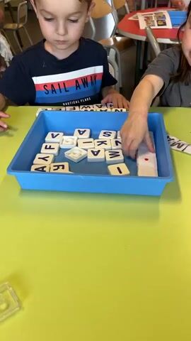 Two young children sit at a bright green table working on an activity involving letter tiles. The children are near each other but appear to be working independently. In front of them is a blue plastic tray filled with white square tiles with black letters on them. The girl, with brown hair, is assembling the tiles to match the names printed on strips of paper that are spread out on the table. The names include "KASSIMA", "MIRA", and "SOHAN." The boy, wearing a "SAIL AWAY" t-shirt, is also using the tiles, though his specific task is not discernible. A small green object is on the table. The room seems to be a classroom, with other children and tables visible in the background. The camera remains mostly stationary, focusing on the children and the letter tiles.