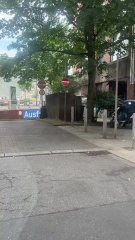 The video shows a view from inside a vehicle, looking out at a street scene in a European city. A large delivery truck with "MÖBEL KUNST" printed on its side is parked on the right side of the street. The truck is white with gold lettering and has a loading ramp at the back. The street is paved with interlocking bricks, and there are several buildings lining the street. The buildings are modern in design, with balconies and large windows. The sky is overcast. There are a few people visible in the video, including a person sitting on a bench outside one of the buildings and a person walking in the distance. The video appears to have been filmed from a moving vehicle, as there is some slight camera shake.