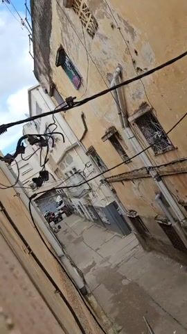 The camera looks down at a narrow alleyway with aging buildings on either side.  The buildings are painted in shades of cream and yellow, with some areas showing peeling paint and water stains. Windows, some with decorative metal bars, are visible on both buildings. A jumble of tangled black electrical wires crisscrosses the alleyway. Down the alley, several vehicles, including a white van and a dark car, are parked.  The pavement is gray and appears to be concrete. The sky above is visible with patches of blue and some clouds. In the background, an indistinct voice speaks.