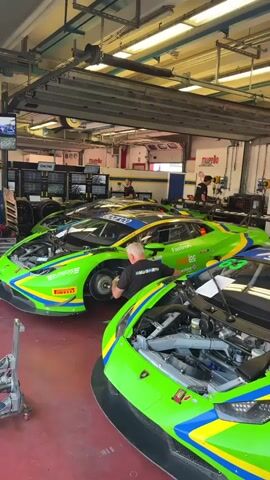 The video shows a pit lane with three race cars. The cars are bright green with yellow and blue accents and have "Pirelli" branding. Two of the cars are Lamborghinis with their hoods open, revealing the engines. Several people are working on the cars. In the foreground, a person in a black shirt is wiping down the front of a Lamborghini. To the left, another person in a black shirt is working on the wheel of a car. In the background, a person in a white shirt is also working on a car. The environment appears to be a garage or workshop, with fluorescent lighting and various tools and equipment visible. The floor is a reddish-brown color.