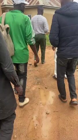 The media file shows a group of people standing on a dirt path in what appears to be an outdoor market setting, possibly Kassa Market in Nigeria. The ground is damp with visible puddles, suggesting recent rain or that the area is naturally moist. In the foreground, a person wearing a black jacket and dark jeans is visible, facing away from the camera. To their left, a person in a bright green long-sleeved shirt and black jeans with a light-colored backpack slung over their shoulder is also seen from behind. Another individual in a striped shirt and green pants is further in the background, also facing away.

The overall atmosphere seems casual, with people gathered. The light suggests it is daytime, though the exact time is difficult to ascertain. The buildings in the background are simple structures with corrugated metal roofs, typical of many market areas. There is no discernible text or signage visible in the frame. The focus is on the individuals and their casual stances, with no clear indication of a specific event or urgent activity.