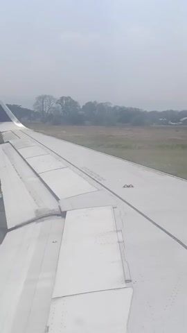 The video shows a view from an airplane window. The airplane wing is visible in the foreground, with flaps extended, suggesting takeoff or landing. The runway is visible to the left, and beyond that, a field of grass and trees are seen under a hazy sky. There are no people visible in the video. The scene appears to be an airport environment.