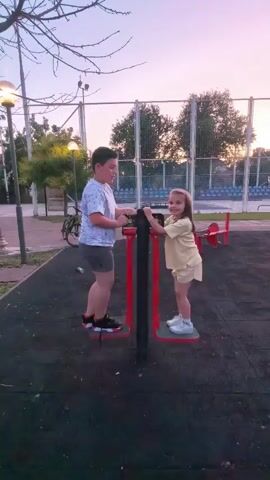 A young boy and girl are exercising on an outdoor fitness machine. The boy, on the left, is wearing a white t-shirt with a graphic design and grey shorts. He has dark hair and is looking at the girl. The girl, on the right, is wearing a yellow t-shirt and yellow shorts. She has long brown hair and is smiling at the boy. They are both standing on pedals of the machine, which has red and black frame. The background shows a park or playground area with trees, a fence, and some athletic equipment. The sky is a pale pink and orange, suggesting dusk or dawn.