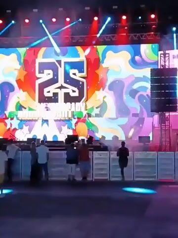 The video shows an outdoor music festival at night. A large stage with a colorful video screen displays the number 25 and the name of the event. Multicolored lights shine down on the stage and the audience. A crowd of people stands in front of the stage. A nearby bar is brightly decorated with a colorful psychedelic pattern. People are standing at the bar, presumably ordering drinks. There are trees and buildings in the background. The video seems to be shot by someone walking through the crowd.