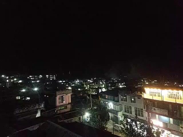 The video shows a nighttime aerial view of a city. Numerous lights illuminate the buildings and streets, creating a sprawling urban landscape against the dark sky. The camera appears to be stationary, capturing the consistent glow of the city.