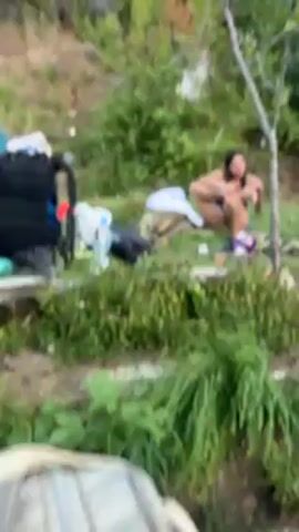 A woman with long dark hair is squatting on the ground in a grassy area, sorting through items. She is wearing a black tank top and dark shorts. Behind her are several backpacks, a tent, and bottles of water. There is a thin tree beside her. The area appears to be a campsite. She smiles and touches her hair. The background is a green hillside with trees and shrubs.
