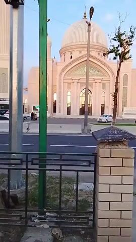 The video shows a view from a moving vehicle, possibly a bus or car, traveling along a road. A white mosque with a large dome and minaret is visible on the right side of the road. The road is divided by a median and has traffic on both sides. A metal railing and brick pillars run along the side of the road closest to the camera. Trees line the sidewalk. The sky is pale blue and the lighting suggests it is early morning or late afternoon. A person is walking on the sidewalk.