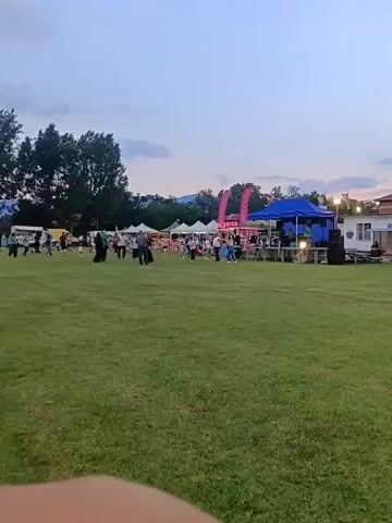 A wide shot of an outdoor event in a grassy field at dusk captures various people moving about. Several white tents, a blue tent, and a pink banner are visible on the right side of the frame, with more tents in the background on the left. Trees line the background, and hills are visible in the distance under a clear sky. A speaker system is set up near the blue tent, and people are gathered around the tents. The grass in the foreground is lush and green.