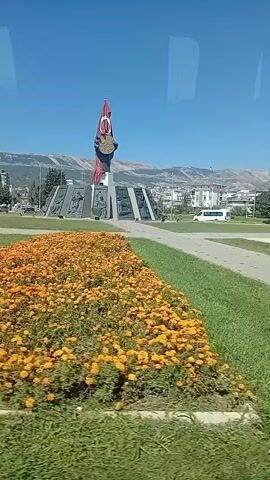 A drone shot pans across a clear blue sky and a green, grassy area with a flower bed of yellow flowers. In the center of the frame, a monument with a large Turkish flag stands tall. The monument is surrounded by a circular plaza with a paved walkway. Buildings and mountains are visible in the background.