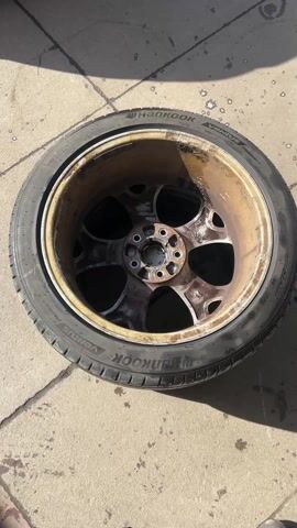 The video shows a close-up view of a dirty car wheel with a tire still mounted. The wheel has a corroded, rusty appearance with traces of old tire sealant around the rim. The tire is black and wet, with the "HANKOOK" brand name visible along the sidewall. A thin layer of bubbly liquid is visible where the tire meets the wheel. The background is a concrete surface, likely outdoors.