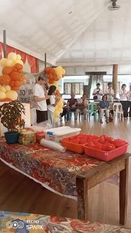 A gathering appears to be taking place under a covered, open-air structure. In the foreground, a long wooden table is laden with items. Two red rectangular baskets overflow with bright red fruit, likely apples or tomatoes, while a clear plastic container with a blue lid sits beside them. A potted plant with dark green leaves is on the left side of the table. The table is covered with a vibrantly patterned tablecloth.

In the mid-ground, two young people, a male with styled blonde hair and a female with dark hair, stand near a display of balloons. The male is wearing a white t-shirt and jeans, and the female is in a white top and blue jeans. They seem to be interacting, possibly presenting something or receiving an award. Behind them, a group of people are seated on chairs, facing the two individuals. Further back, another group of people stand, many wearing white shirts with lanyards around their necks, suggesting some kind of organized event or presentation. One person in this standing group is holding a microphone.

The scene is brightly lit, suggesting it's daytime. The ceiling is white and appears to be corrugated material. A ceiling fan is visible overhead. The overall atmosphere seems festive, with the balloons and the gathering of people. The presence of a tablecloth with a Philippine flag motif and the "TECNO SPARK" watermark in the corner suggests the location is the Philippines, potentially Dumaguete as indicated by the prompt. The text "SO MUCH SERVICE" is visible on a clear plastic banner behind the two main subjects. The event seems to be a celebration or recognition ceremony.