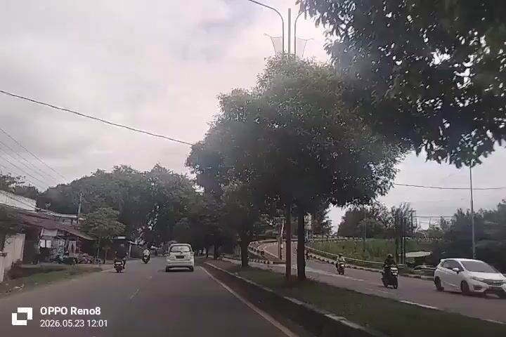 The video shows a street view from inside a moving vehicle. There is a car driving on a road lined with trees. On the left side of the road, there are shops and some people walking around. On the right side of the road, there is a highway overpass. A few motorcycles and cars are seen driving on the road alongside the vehicle filming. The weather appears to be overcast. The camera is an OPPO Reno8 and the timestamp indicates 2026.05.23 12:01.