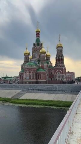 A vertical video pans slightly left across a large, ornate red and green cathedral with golden domes. The cathedral is set against a cloudy sky. In the foreground, a body of dark water is visible, with green vegetation along its bank. A concrete embankment with a metal railing runs between the water and the cathedral. A few people are seen walking on the embankment and stairs leading down to the water.