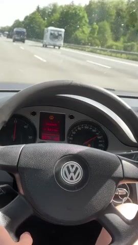 The view from inside a moving car shows the dashboard and steering wheel. The car appears to be a Volkswagen. The dashboard lights are on and display information in German. The car is driving on a highway or freeway with other vehicles visible in front. The road is asphalt with lane markings. The weather appears to be sunny or partly cloudy. Trees are visible on the side of the road. The driver's hands are visible on the steering wheel.