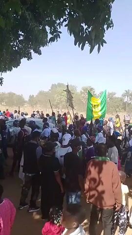 A large crowd of people has gathered in an outdoor, possibly rural setting. There is a car in the midst of the crowd. Some individuals are holding flags and what appear to be ceremonial staffs or poles. The flag is green and yellow with a symbol. People are dressed in a variety of clothing, including casual wear and more traditional attire. The weather appears to be sunny, and the surroundings include trees and open fields.