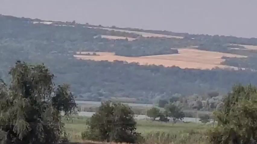 A slow pan right across a rural landscape. In the foreground, there are dry, brown grasses and some green reeds. Further back, there is a body of water, possibly a river, with trees and bushes lining its banks. In the background, there are rolling hills covered in trees and fields. The sky is overcast. There are no people visible in the video.