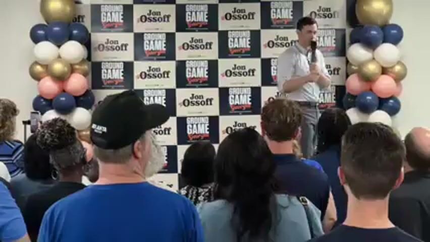 A man identified as Jon Ossoff is giving a speech to a seated audience at an indoor event. He stands before a backdrop featuring repeated "GROUND GAME Georgia" and "Jon Ossoff" logos. Balloon arrangements in gold, white, pink, and navy blue frame the backdrop on both sides. Ossoff is wearing a light-colored button-down shirt and dark trousers, holding a microphone in his right hand and gesturing with his left. The audience, diverse in age and ethnicity, is seen from behind, facing the speaker. One audience member wears a black baseball cap with "ATLANTA UNITED" visible on it. The atmosphere appears to be a political rally or campaign event in Decatur, United States.