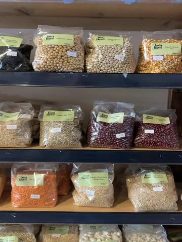 The video shows a close-up, eye-level shot of shelves filled with various dried beans, lentils, and grains packaged in clear plastic bags. Each bag has a yellow label with green text. The products are arranged in rows on dark shelves, and the overall environment appears to be a store or market. The lighting is bright and even. There are no people visible in the video. The camera slowly pans across the shelves, allowing a view of different types of legumes and grains.