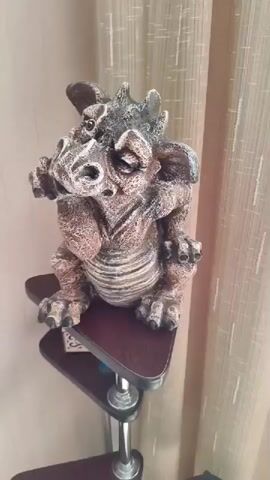 A decorative dragon figurine is displayed on a multi-tiered shelf. The figurine is brown and gray with intricate detailing, depicting a dragon with wings, horns, and claws. It is positioned on the top tier of the shelf, which is a triangular wooden platform. Below this, two more triangular shelves are visible, also made of wood and supported by a silver metal pole. The background features beige curtains, suggesting an indoor setting. A green spray bottle is visible on the lowest shelf. The camera slowly zooms out, providing a wider view of the figurine and its surroundings.