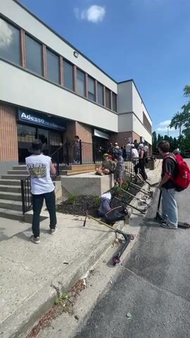 A group of young people are gathered outside a brick and white building on a sunny day. Several of them have scooters, and some appear to be preparing to ride or are in the process of riding them. One individual is standing on a scooter near the entrance of the building, which has stairs leading up to it. Another person in a white tank top and dark pants with a cowboy hat is holding a black strap and appears to be observing the activity. In the background, other individuals are seated or standing near the entrance, with scooters parked nearby. The building has signage that reads "Adesso" and "DINE-IN TAKEOUT CATERING." The ground is paved with concrete, and there is a curb and a street visible in the foreground. Some vegetation is growing along the edge of the concrete.