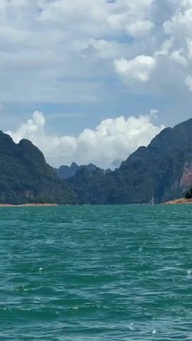 The video shows a view from a body of water, likely a lake or a large river, looking towards a mountainous landscape. The water is a deep turquoise green with small waves and ripples, indicating some movement. In the background, several large, lush green mountains rise steeply from the water's edge. The mountains are covered in dense vegetation, with some rocky outcrops visible. The sky above is a bright blue, dotted with fluffy white clouds. The overall impression is of a scenic, natural environment, possibly in a tropical or subtropical region. There are no people or man-made objects clearly visible in the frame.