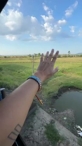 The video shows a person's arm and hand extended out of a vehicle window, with the hand open and fingers spread, reaching towards the sky. The arm has tattoos on the forearm and is adorned with bracelets. The background is a landscape of green fields, possibly rice paddies, under a bright blue sky with white clouds. A utility pole with wires is visible in the distance. The camera appears to be mounted inside the vehicle, looking out. The video is short, lasting only a few seconds, and the scene is captured in daylight.