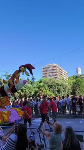 This is a vertical, outdoor video taken on a sunny day with a clear blue sky. The foreground shows the back of several people's heads and shoulders as they look towards a parade float. The float is elaborately decorated with flowers and depicts characters from Looney Tunes. Specifically, Wile E. Coyote is shown riding a rocket, and the Road Runner is perched atop a cactus. The float also has a sign that says "Beep Beep." The float is surrounded by a crowd of spectators, many of whom are holding up their phones to take pictures or record video. Some people in the crowd are wearing red shirts and appear to be part of the parade, perhaps participants or organizers. In the background, there are trees and a multi-story building. The overall scene suggests a festive atmosphere, likely a parade or festival.