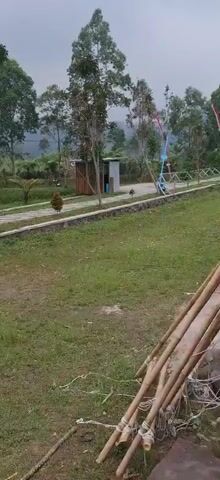 The video shows an outdoor scene with a paved walkway winding through a grassy area with trees and plants. In the background, there is a building with a traditional roof and several tents set up. A flag with red and white stripes is planted on a pole. People are walking on the path and near the tents. The environment appears to be a park or a campsite with hilly terrain and lush greenery.