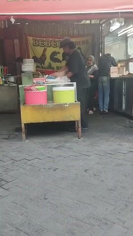 A street food stall is shown. The stall has a banner that reads "BEBEK GORENG BAKAR". There are three people visible, all wearing dark clothing. One person is standing behind a counter, interacting with another person who is on the other side of the counter. A third person is further down the counter, also appearing to be working. In front of the counter are two large, colorful containers, one pink and one green. The ground is paved with grey bricks.