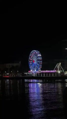 The video shows a Ferris wheel at night, illuminated with red, white, and blue lights. The Ferris wheel is situated on a pier or boardwalk, with a body of water in the foreground. The water reflects the lights of the Ferris wheel and other amusement park attractions, creating a colorful display. There are also other amusement rides visible in the background, as well as buildings and trees along the shoreline. The overall scene is a festive and lively atmosphere of a nighttime amusement park.