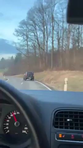 The video shows the view from inside a car as it travels along a rural road. The dashboard and steering wheel of the car, a Volkswagen, are visible. The car is traveling on a two-lane road with a grassy median on either side. There are trees and fields visible in the background under a cloudy sky. Ahead of the car, another vehicle, an older model black Jeep, is also traveling along the road. The car maintains a consistent following distance from the Jeep.